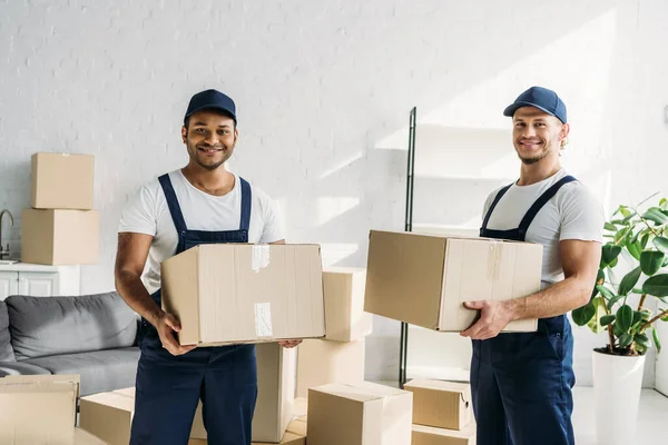 Cheerful Multiethnic Movers Caps Uniform Carrying Boxes Apartment — Stock Photo, Image