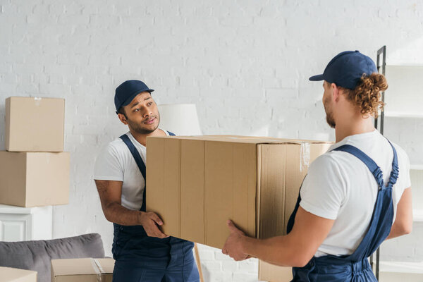 young multiethnic movers in uniform carrying huge carton box 