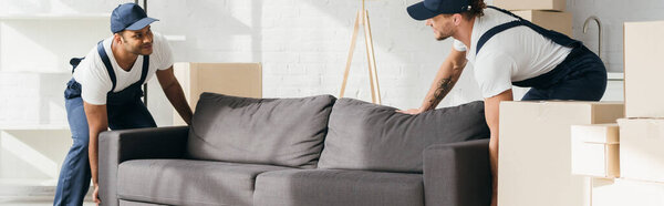 happy multicultural movers in uniform smiling while carrying couch in apartment, banner