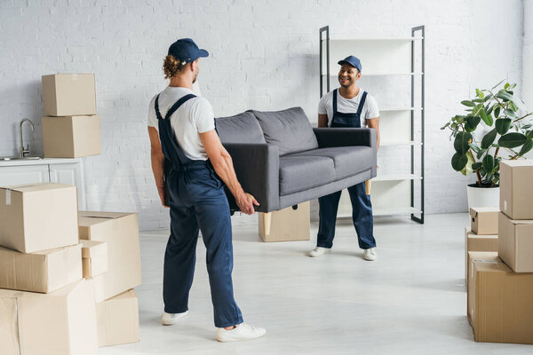 full length of happy young multiethnic movers in uniform carrying couch in apartment