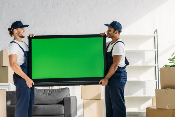 side view of happy multicultural movers in uniform carrying plasma tv with green screen in apartment