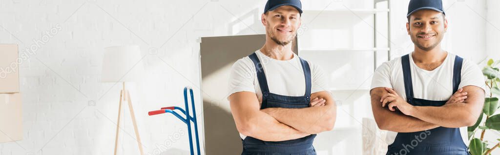 cheerful multicultural movers in caps standing with crossed arms in apartment, banner