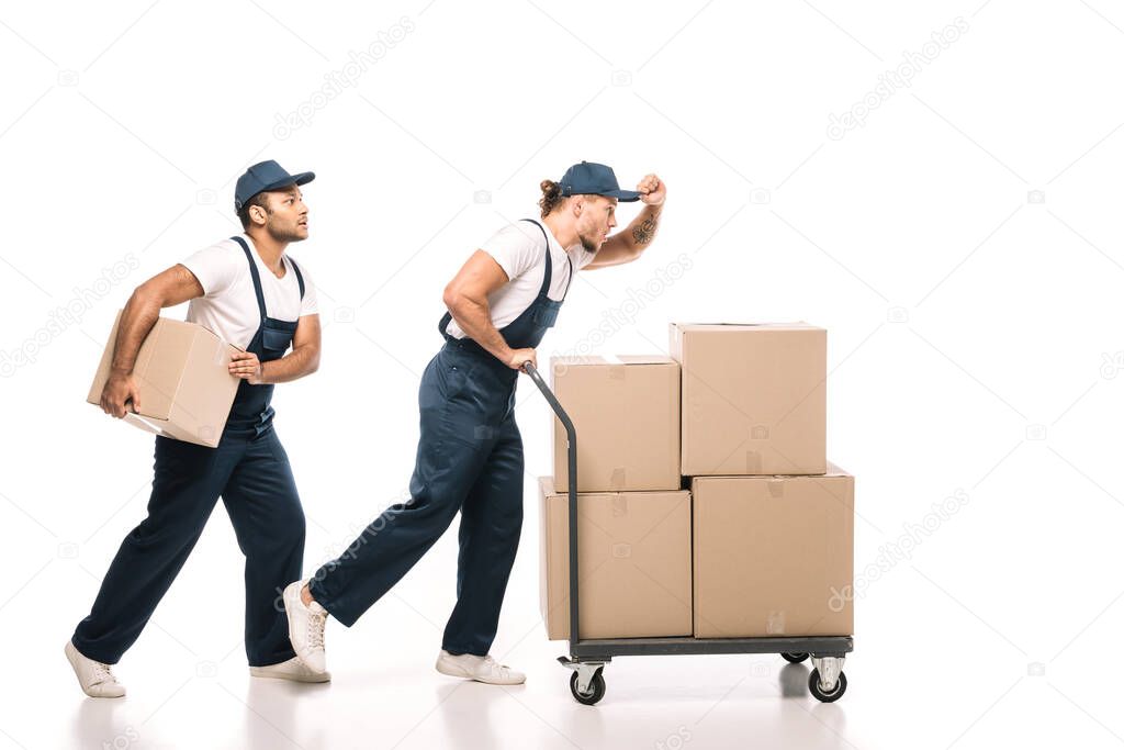 full length of multicultural movers in uniform and caps walking near hand truck with carton boxes on white