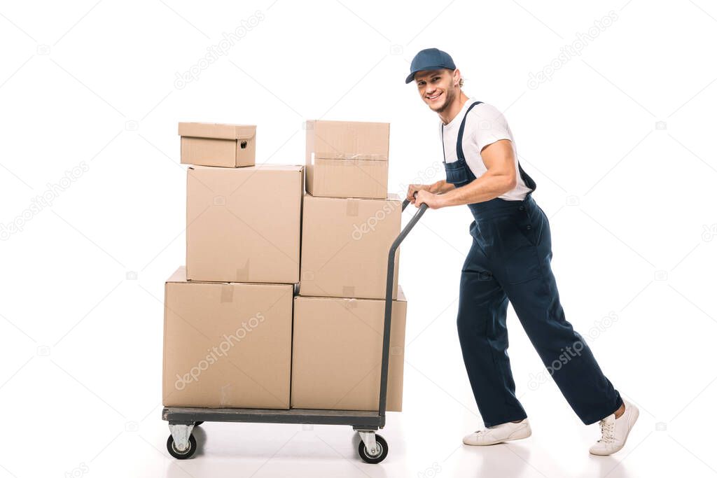 full length of cheerful mover in uniform and cap pulling hand truck with carton boxes on white