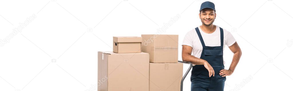 cheerful indian mover in overalls standing with hand on hip near hand truck with carton boxes isolated on white, banner