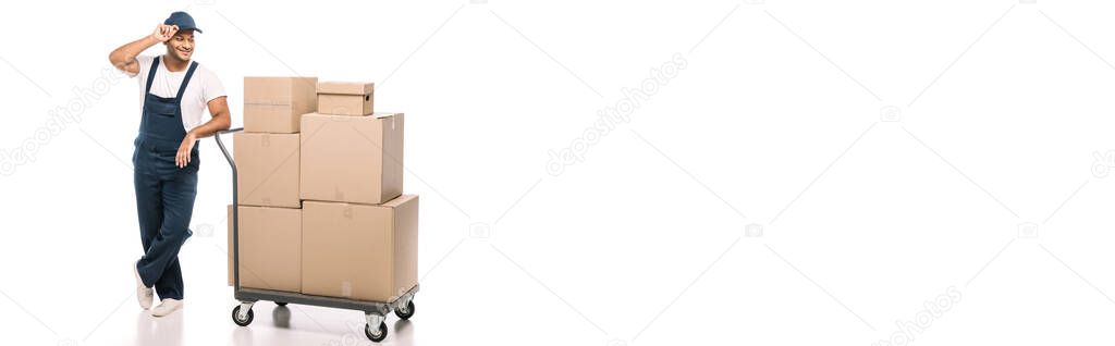 full length of joyful indian mover standing near hand truck with carton boxes and adjusting cap on white, banner