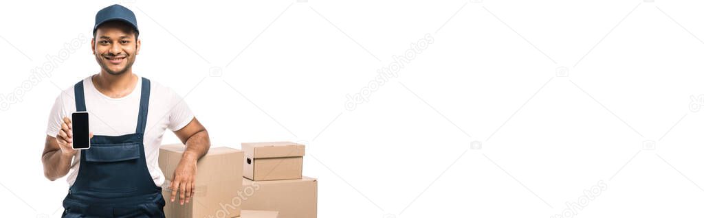 happy indian mover in overalls holding smartphone with blank screen near carton boxes isolated on white, banner