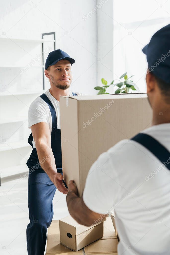 young worker in uniform receiving carton box from indian coworker on blurred foreground