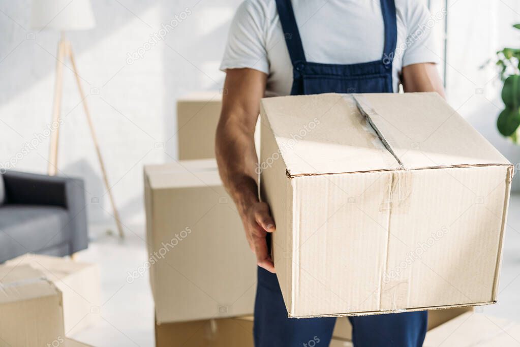 partial view of mover in uniform holding carton box in apartment 
