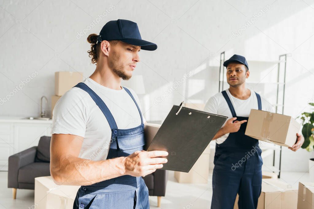 man in cap and uniform looking at clipboard near indian coworker pointing with finger at box on blurred background 