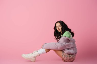 Pembe arka planda yerde poz veren şık kışlık kıyafetli esmer bir kadının yan görüntüsü