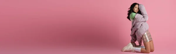 側ビューのブルネット若い女性でスタイリッシュな冬服で膝の上にポーズピンクの背景 バナー — ストック写真