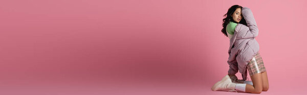 вид сбоку брюнетки молодая женщина в стильном зимнем наряде позируя на коленях на розовом фоне, баннер