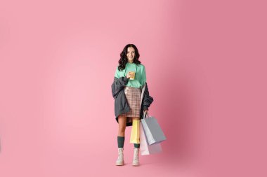 Gülümseyen esmer, günlük kışlık kıyafetli, alışveriş torbaları ve pembe arka planda kahve olan genç bir kadın.