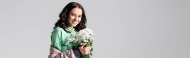 smiling brunette young woman in stylish winter outfit posing with flowers on grey background, banner
