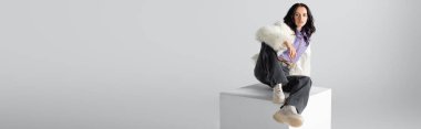 elegant brunette young woman in stylish faux fur jacket posing on cube on white background, banner clipart