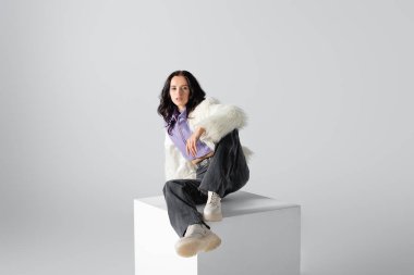elegant brunette young woman in stylish faux fur jacket posing on cube on white background clipart
