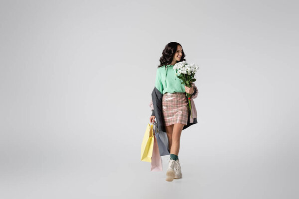 улыбающаяся брюнетка молодая женщина в стильном зимнем наряде позирует с цветами и сумками для покупок на сером фоне