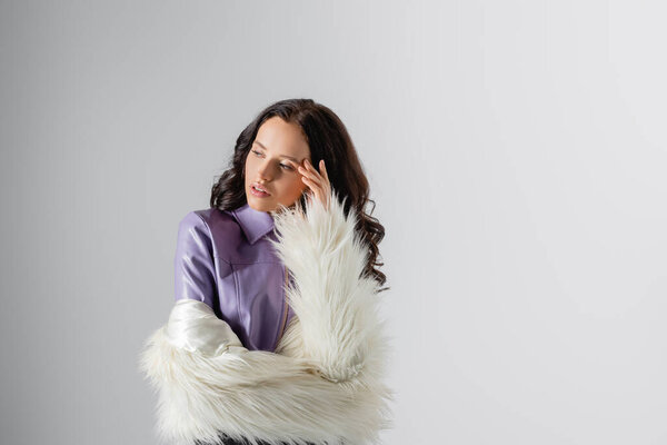 brunette young woman in stylish white faux fur jacket posing on grey background