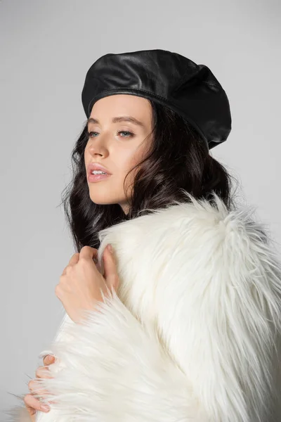 エレガントなブルネット若いです女性でスタイリッシュなホワイトフェイクファージャケットと革ベレー帽ポージング上の灰色の背景 — ストック写真