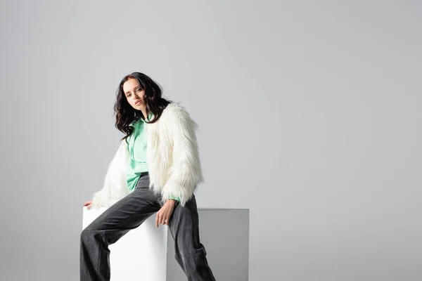 brunette young woman in faux fur jacket posing on cube on white background