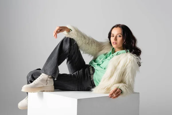 brunette young woman in faux fur jacket posing on cube on white background