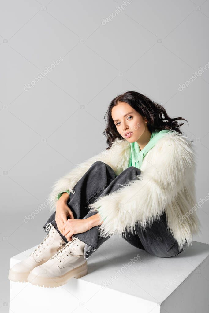 brunette young woman in faux fur jacket posing on cube on white background