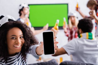 Mutlu Afro-Amerikalı kadın, arka planda bulanık arka planda çok kültürlü futbol hayranlarının yanında boş ekranlı akıllı telefon gösteriyor.