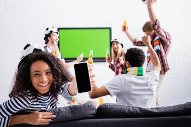 Neşeli Afro-Amerikalı kadın, arka planda bulanık şapkalarla futbol hayranlarının yanında boş ekranlı akıllı telefon gösteriyor.