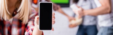 Arkaplanı bulanık arkadaşlarının yanında elinde boş ekranlı akıllı telefon tutan bir kadın görüntüsü, afiş