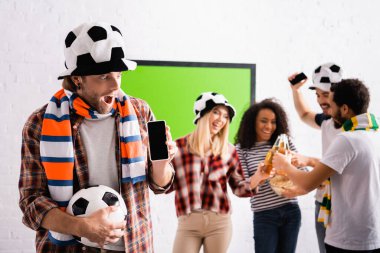 heyecanlı futbol fanatiği futbol topu tutuyor ve arka planda bulanık arka planda bira tokuşturan çoklu etnik arkadaşların yanında boş ekranlı akıllı telefon.