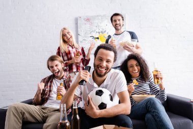 excited african american man with soccer ball watching football championship with multicultural friends showing win gesture clipart