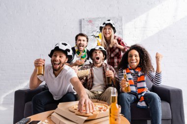 excited man taking pizza while watching football championship with multicultural friends on blurred foreground clipart