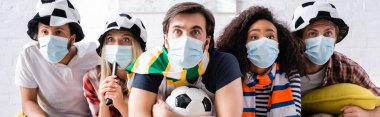 worried multiethnic friends in football fans hats and medical masks watching championship at home, banner clipart