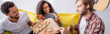 Çok kültürlü adamların yanında elinde pizza ve pankart tutan akıllı telefonlu neşeli Afrikalı Amerikalı kadın.