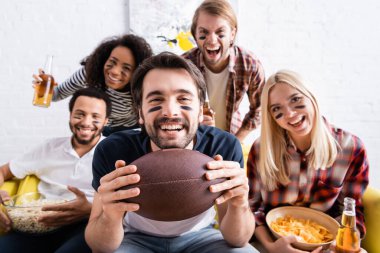 Heyecanlı adam rugby topu tutuyor ve arka planı bulanık renkli çok ırklı arkadaşlarının yanında şampiyonluğu seyrediyor.
