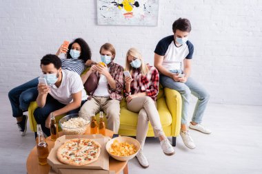 Tıbbi maskeli çok kültürlü dostların abur cuburların yanında akıllı telefonlardan sohbet ederken ve masada bira içerken yüksek açılı görüntüsü.