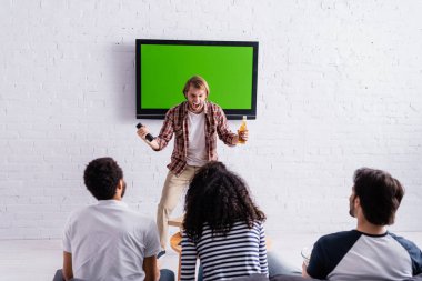 Heyecanlı adam bağırıyor ve lcd TV 'nin yanında jest yapıyor. Bu arada çok kültürlü arkadaşlar spor müsabakalarını izliyorlar.