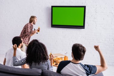 Heyecanlı adam televizyon başında çığlık atarken, çok ırklı spor hayranları jest yapıyor.