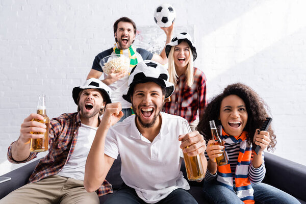 excited multiethnic friends in football fans hats shouting while watching competition at home