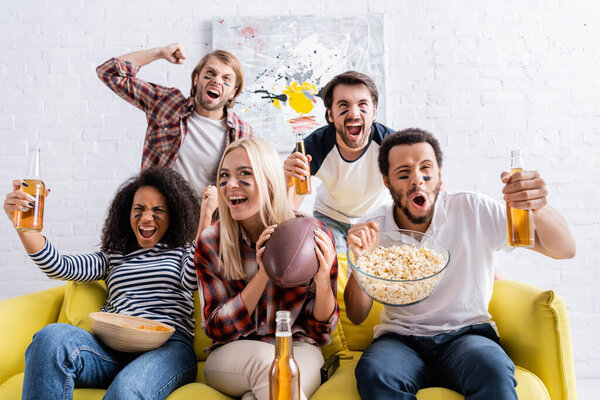 excited multiethnic friends with painted faces holding beer and shouting while watching rugby championship 