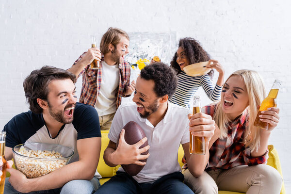 cheerful multiethnic rugby fans with painted faces holding beer while watching competition at home