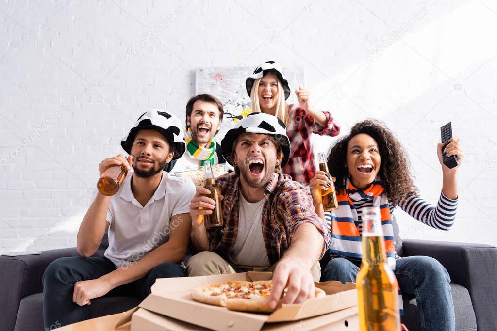 excited man shouting, holding beer and taking pizza while watching championship with multicultural friends on blurred foreground