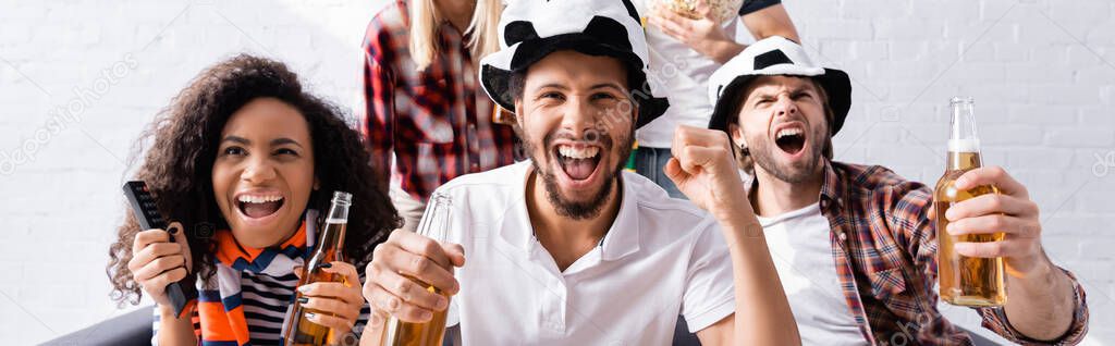 excited man screaming and showing win gesture while watching football competition with multiethnic friends on blurred background, banner