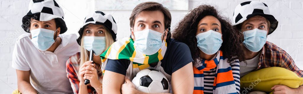 worried multiethnic friends in football fans hats and medical masks watching championship at home, banner