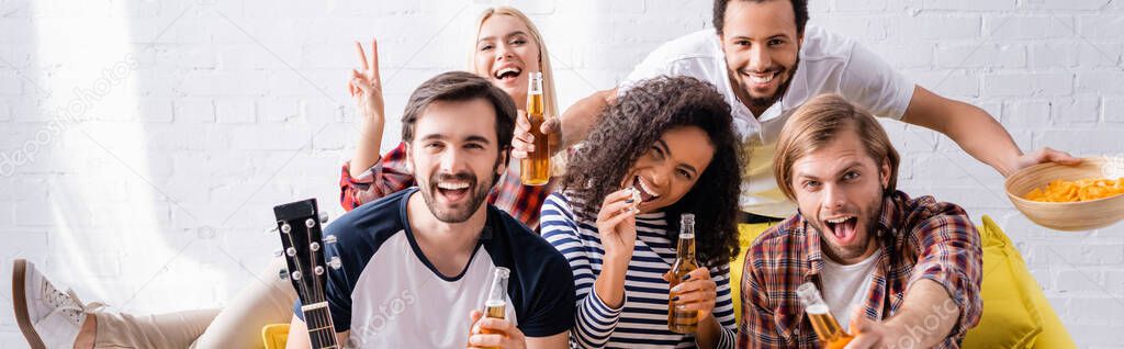 smiling woman showing victory gesture near excited friends holding beer during party, banner
