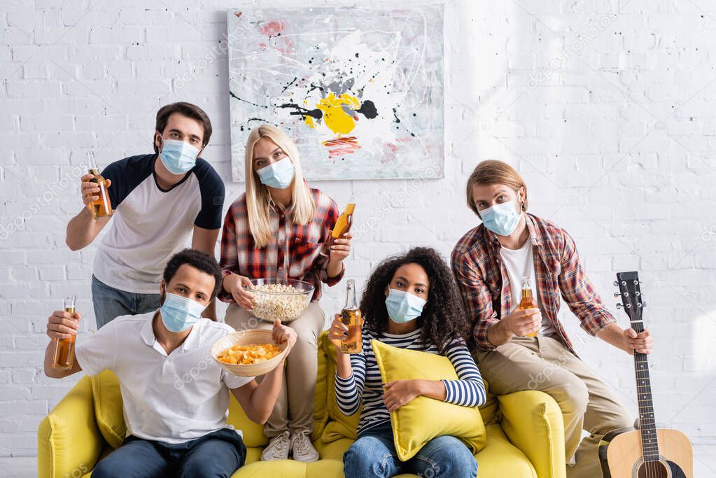 multiethnic friends in medical masks looking at camera while holding beer and snacks during party