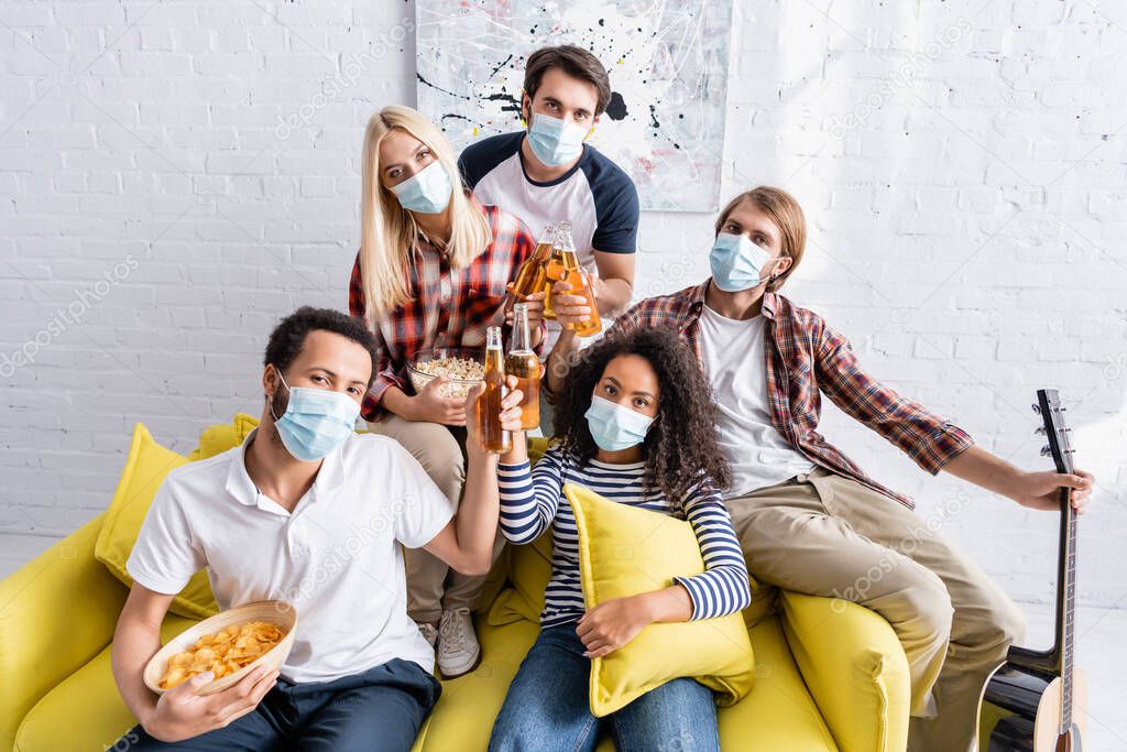 multiethnic friends in medical masks looking at camera while clinking bottles of beer during party