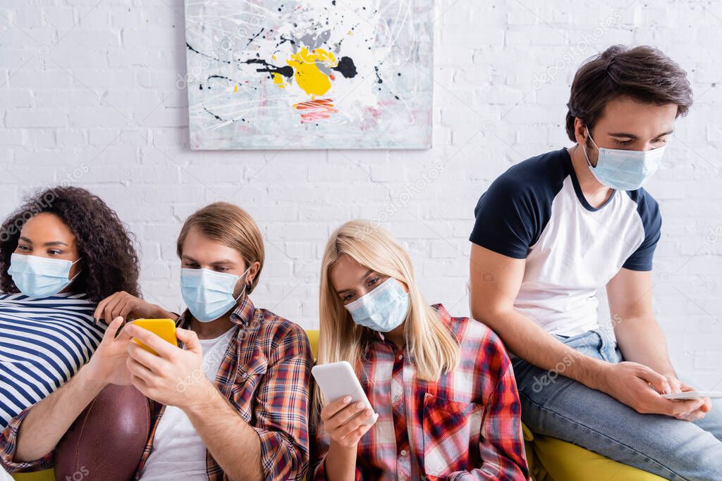 multiethnic friends in medical masks messaging on mobile phones while sitting on sofa