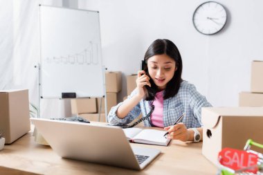 Bulanık ön plandaki yardım merkezinde laptopun yanında telefonla konuşan pozitif Asyalı gönüllü ve karton kutular.